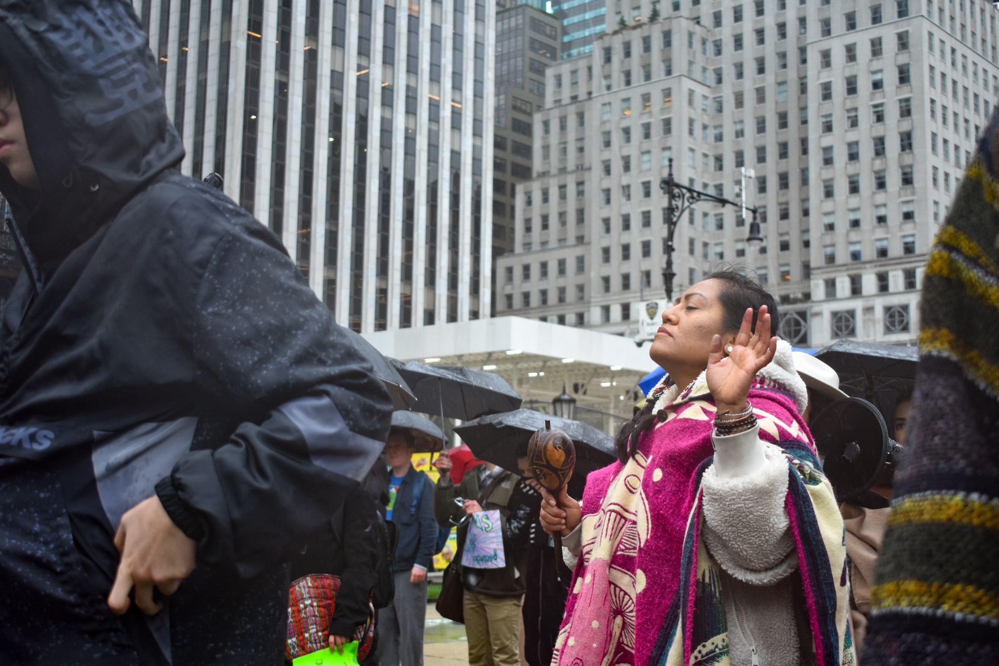 A woman wearing a wool pink and white poncho with a mushroom looks upwards, with her hands outward, with skyscrapers in the distance. 