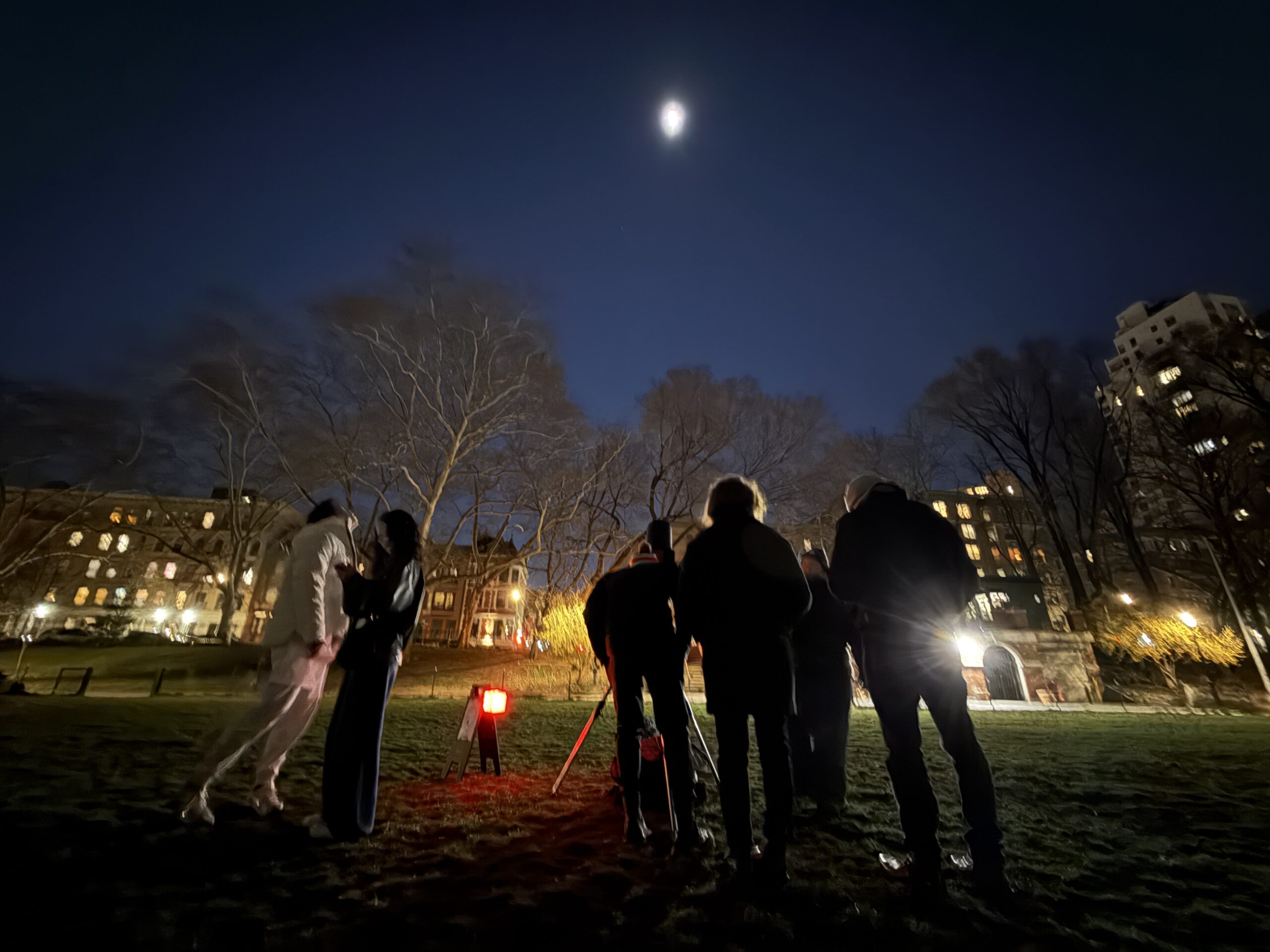 A group of people in a park huddle around a telescope and look at the moon.